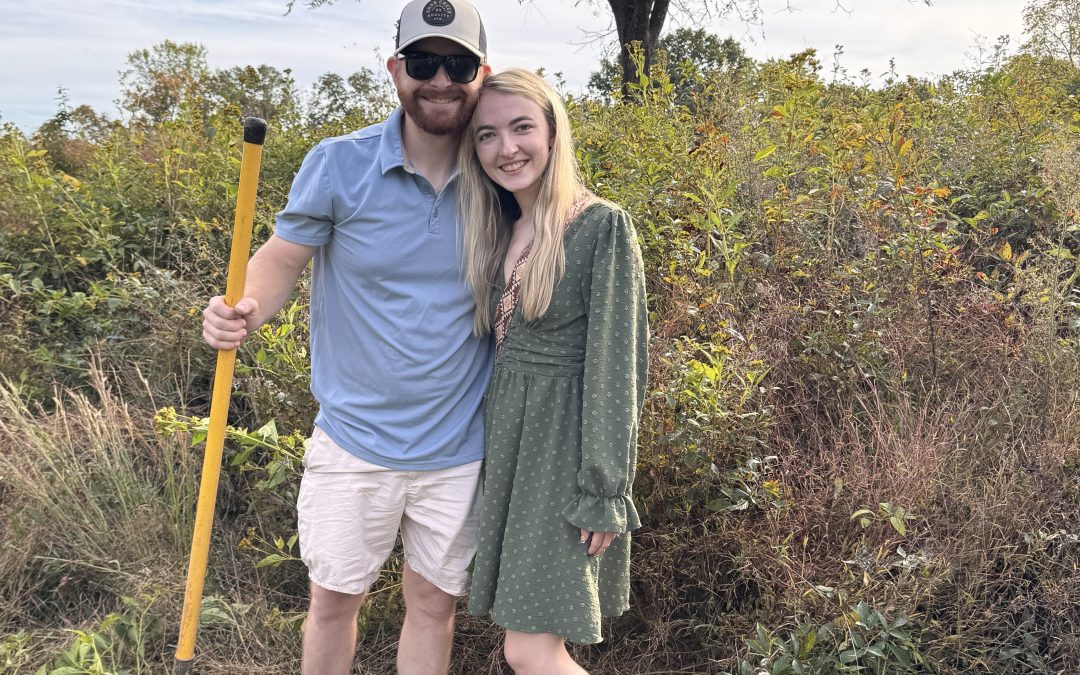 A couple standing together outdoors, one holding a shovel, with greenery in the background.