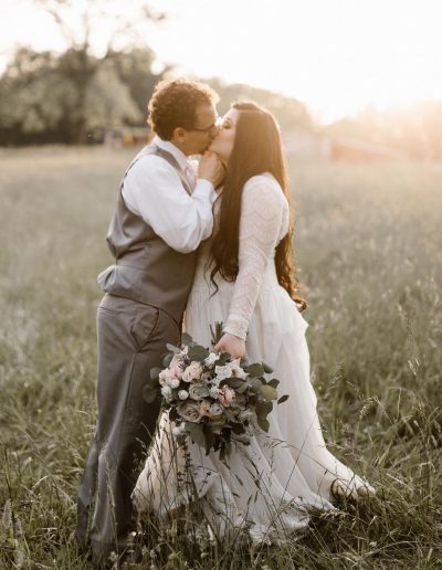 Newlywed couple kisses in a sunlit meadow as the bride holds a pastel floral bouquet beside her groom.