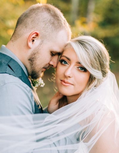 Bride and groom embrace in a soft outdoor portrait, with the bride’s sheer veil flowing around them and warm green bokeh behind them.
