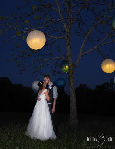 A bride and groom kiss beneath glowing paper lanterns on a tree at dusk, with a deep blue sky and meadow grass surrounding them.
