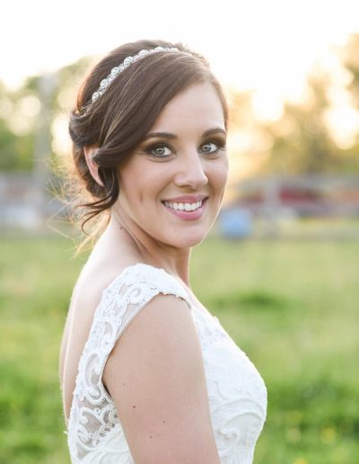 Smiling bride in a lace wedding gown and jeweled headband stands in a sunlit meadow at golden hour with blurred green trees and warm light.