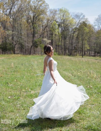 Smiling bride in a flowing white gown twirls in a sunlit meadow with spring trees and soft blue sky at Heartland Meadows wedding venue setting.