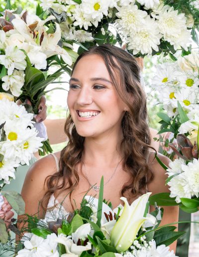 Smiling bride with wavy brown hair holds a bouquet of white flowers beneath an abundant floral arch at a wedding venue