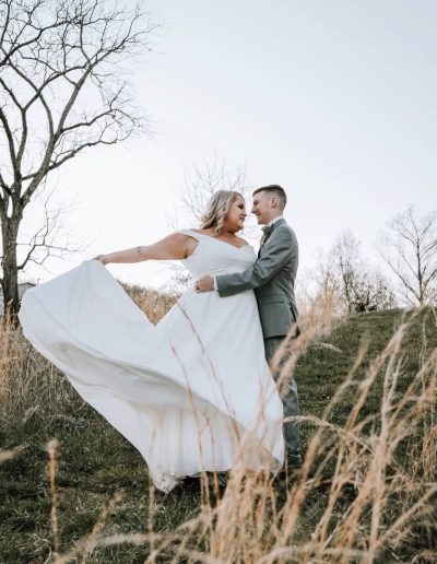 A newlywed couple embraces in a grassy meadow as the bride’s flowing white dress moves beside a leafless tree.