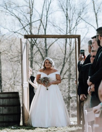 A bride in a white wedding gown holds a bouquet while walking through an ornate doorway toward waiting guests outdoors.