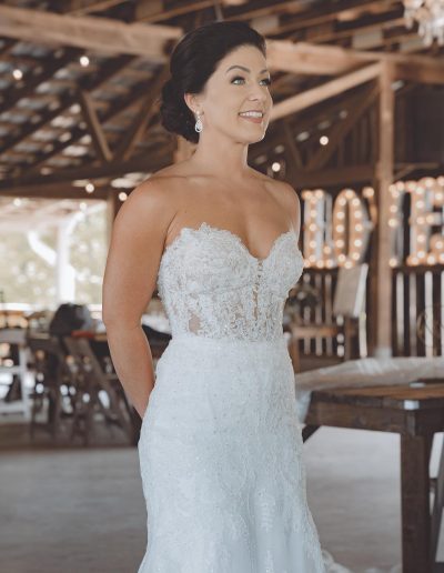 Bride wearing a lace wedding dress with a fitted bodice and a sweetheart neckline, smiling in a rustic venue.