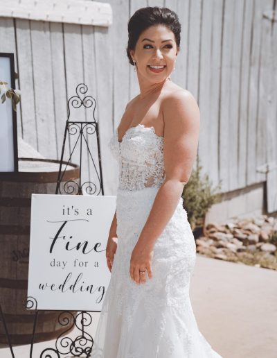 Bride wearing a lace wedding dress standing next to a sign that reads 'it's a Fine day for a wedding'.