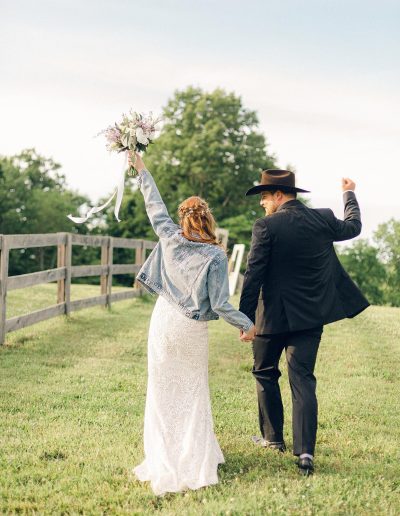 Newlywed couple walks across a grassy meadow, the bride raising her bouquet while the groom lifts his hat beside a wooden fence.