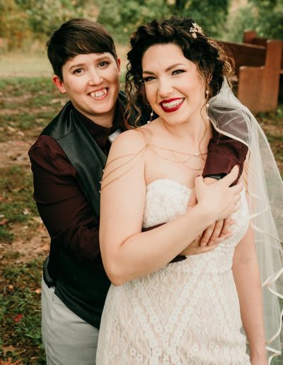 Two individuals in wedding attire, one wearing a white dress and the other in a dark vest, posing together outdoors.