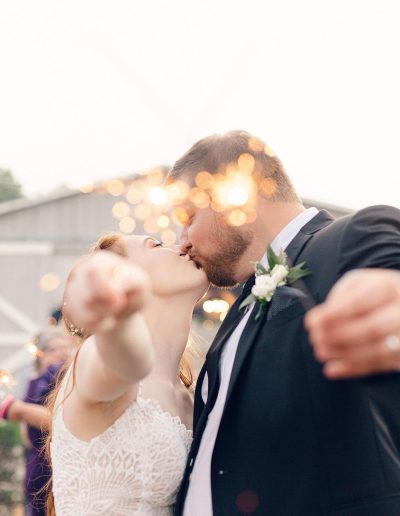 Newlywed couple kisses during an outdoor wedding celebration as guests and warm sparklers fill the background.