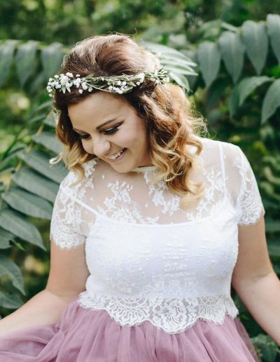Woman wearing a floral crown and a lace top, smiling while standing among green foliage.