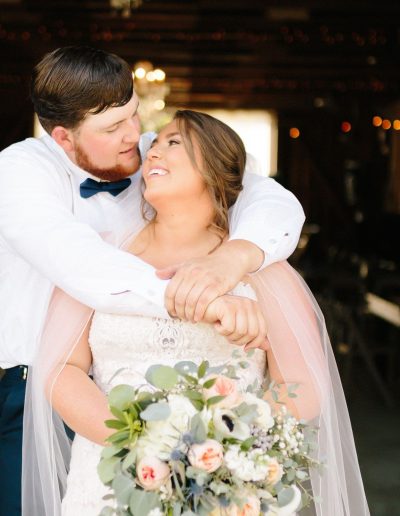 A bride and groom embrace beneath warm string lights in a barn, while she holds a pastel bouquet and they share a tender wedding moment.