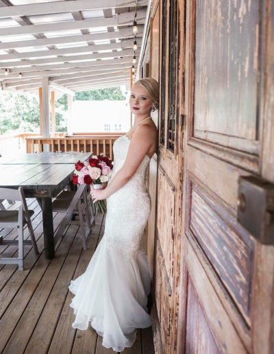 Blonde bride in a white lace mermaid gown holding a red and pink bouquet beside a rustic wooden wall on a covered porch at Heartland Meadows.