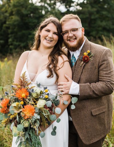 Bride and groom posing together outdoors, holding a floral bouquet with orange flowers and greenery.
