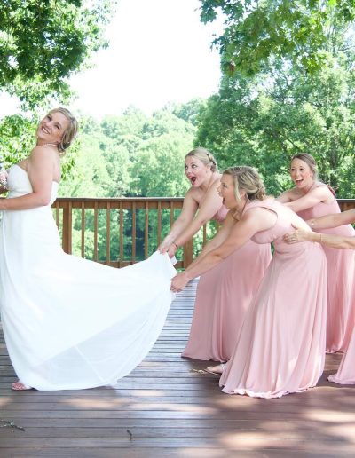 A bride in a white gown laughs as five bridesmaids in pink dresses pull her veil on a sunlit wooden deck among trees.