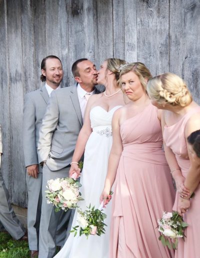 A bride and groom kiss among their wedding party, with bridesmaids in pink dresses and bouquets beside a rustic wooden wall.