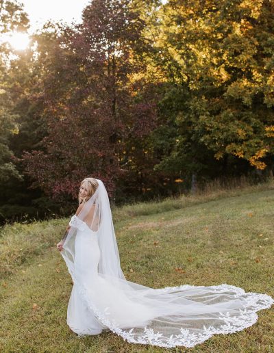 Bride in a white lace gown and long veil stands on a grassy hillside beneath golden sunset light and autumn trees at Heartland Meadows wedding venue setting.