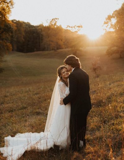 Bride in a lace gown and groom in a black suit embrace in a sunlit meadow at golden hour, with warm autumn tones and a flowing veil.