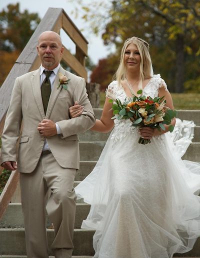 A bride in a white lace gown walks beside a groom in a beige suit along outdoor wedding venue steps.