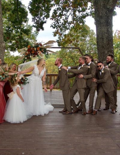 A bride and groom celebrate with bridesmaids and groomsmen on a wooded wedding venue deck beneath mature trees.