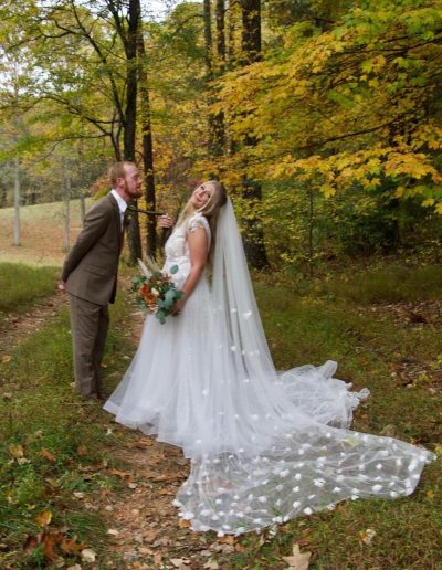 A bride and groom pose on a wooded path, with her floral bouquet, long veil, and beaded train among autumn leaves.