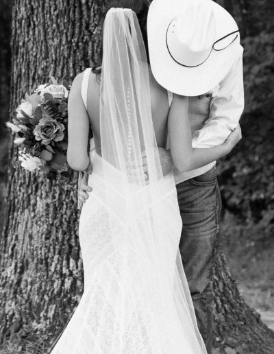 Black-and-white bride and groom embrace beneath a large tree, with a flowing veil, lace gown, bouquet of roses, and a rustic outdoor setting.