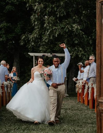 Newlyweds walk down a grassy outdoor aisle, bride in a white gown holding pink flowers while guests cheer beside rustic wooden pews under tall trees.