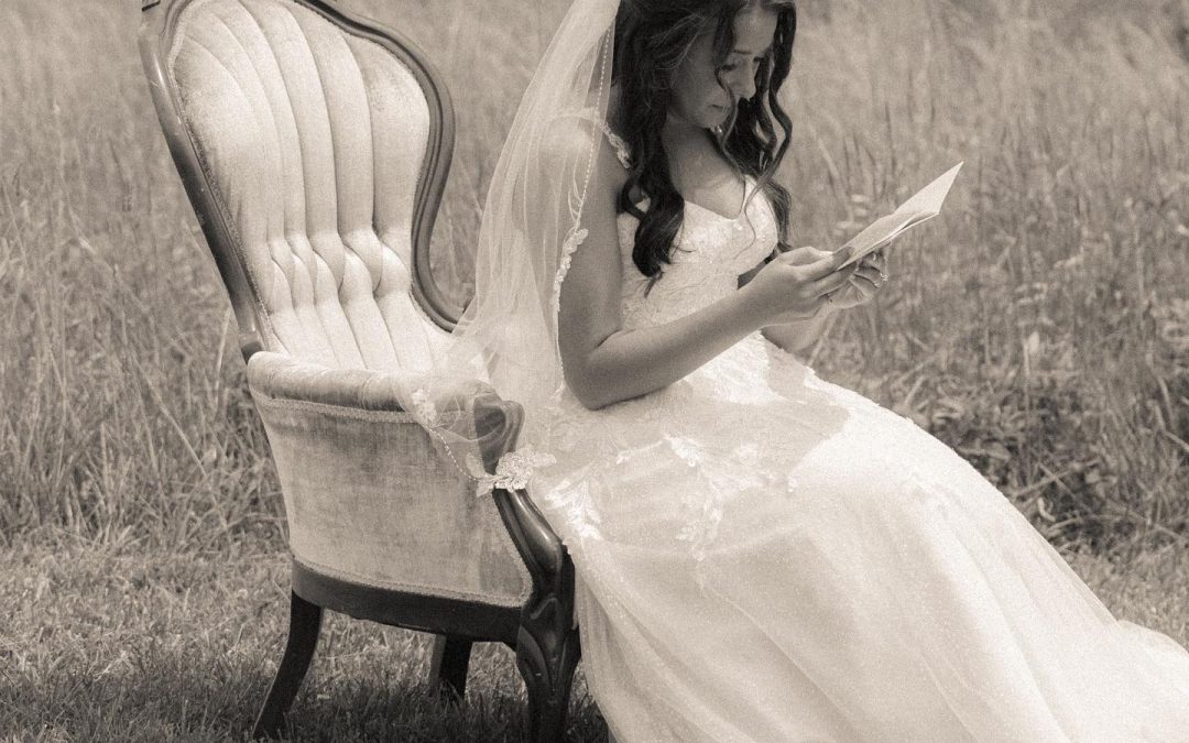 Bride in a white wedding dress sitting on an ornate chair while reading a letter outdoors.