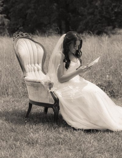 Bride in a white wedding dress sitting on an ornate chair while reading a letter outdoors.