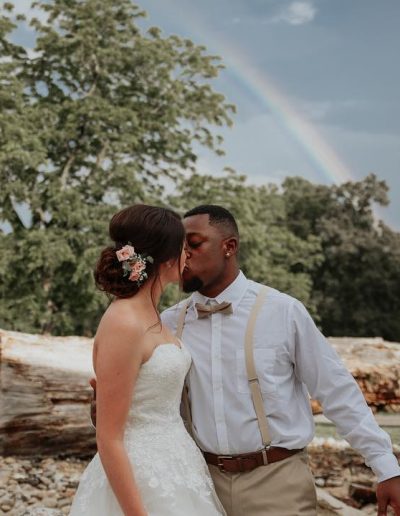 A newlywed couple shares a kiss beneath a soft rainbow, with a white wedding dress, suspenders, and rustic stone landscaping behind them.