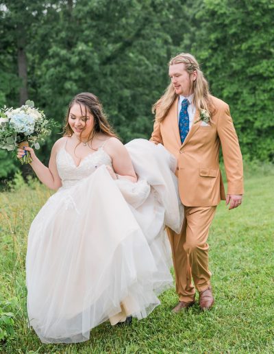 Bride in a white wedding dress and groom in a tan suit walking on a grassy area holding hands and a bouquet.