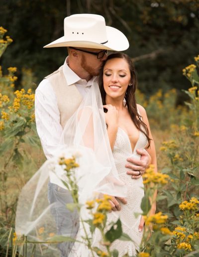 A romantic bride and groom embrace in a wildflower meadow, with a white cowboy hat, lace gown, and soft yellow blooms at Heartland Meadows.
