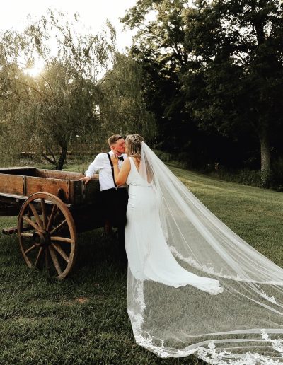 Bride in a white wedding dress and veil with groom near a wooden wagon outdoors.