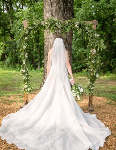 A bride in a flowing white wedding gown stands beneath a rustic wooden arbor draped with greenery in a forested meadow.