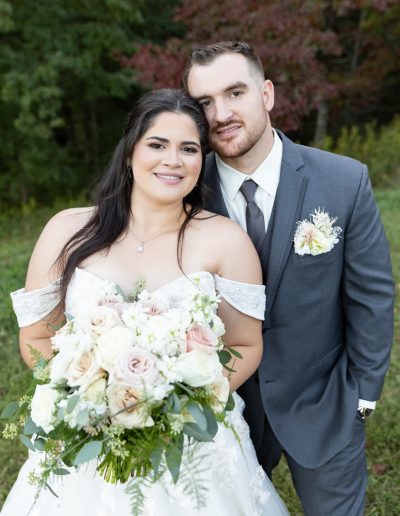 A smiling bride and groom pose outdoors, with the bride holding a lush white and blush floral bouquet.