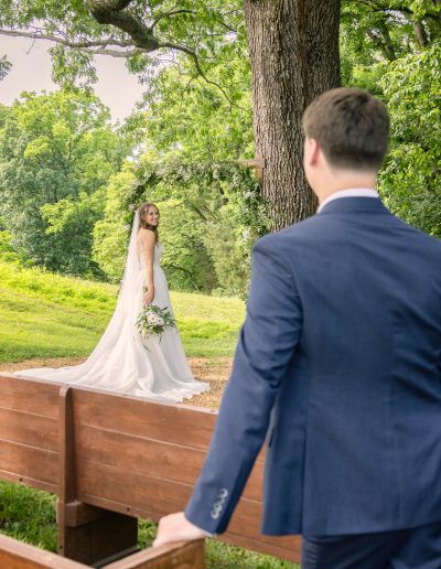 A groom in a navy suit approaches his bride in a white wedding dress beneath a large tree in a lush meadow.