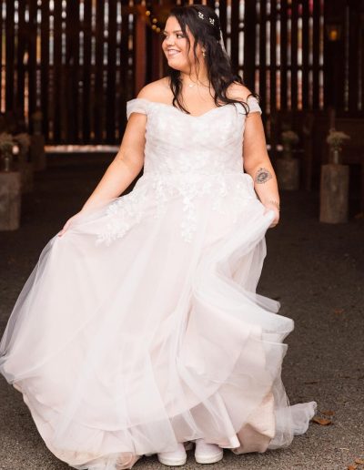 Bride in a flowing white wedding gown smiles inside a rustic barn aisle, with soft drapery, wood beams, and warm string lights behind her.