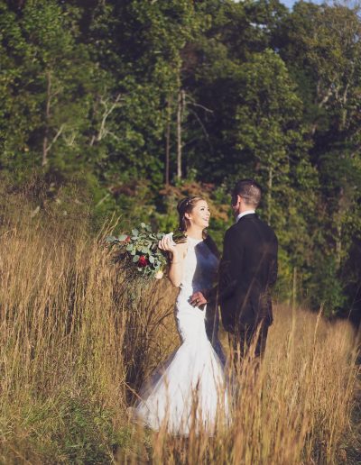Bride in a white wedding dress holding a bouquet, standing with a groom in a suit in a grassy field.