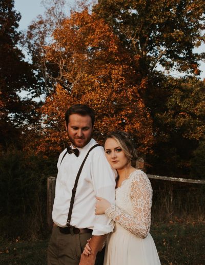 Autumn wedding couple standing close outdoors, with the bride in a lace gown and the groom in suspenders beneath warm orange fall foliage.