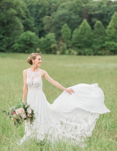 A bride in a white lace gown stands in a grassy meadow, holding a bouquet while her dress flows against a backdrop of lush green trees.
