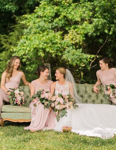 Bride in white veil sits with three bridesmaids in blush dresses on a vintage sofa, holding bouquets beneath leafy trees at Heartland Meadows.