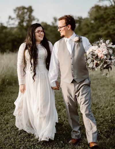 Newlyweds in white and gray wedding attire walk hand in hand through a grassy field, carrying a blush and eucalyptus bouquet at sunset.