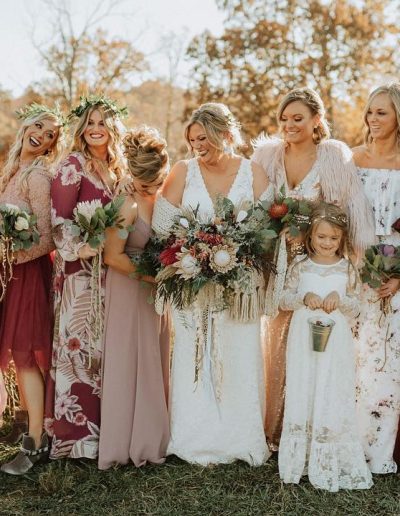 Group of bridesmaids in various dresses holding bouquets, posing outdoors in a natural setting.