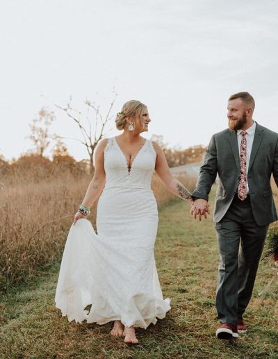 Bride in a white lace wedding dress and groom in a gray suit walking hand in hand with a dog on a grassy path.