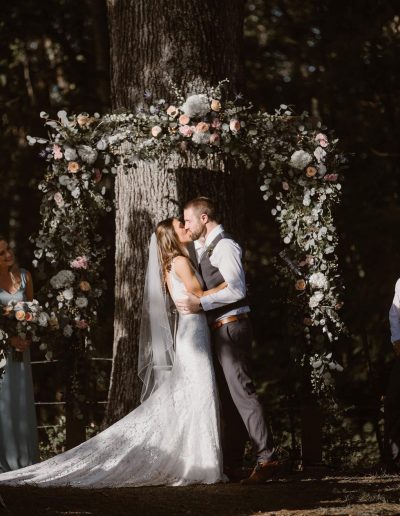 Newlyweds kiss beneath a lush floral arch around a tree, with the bride in a long white gown and groom in a gray suit.