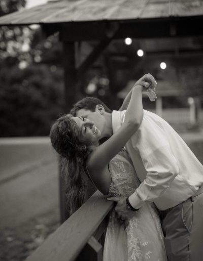 A couple embracing and smiling while leaning against a wooden railing at a wedding venue.