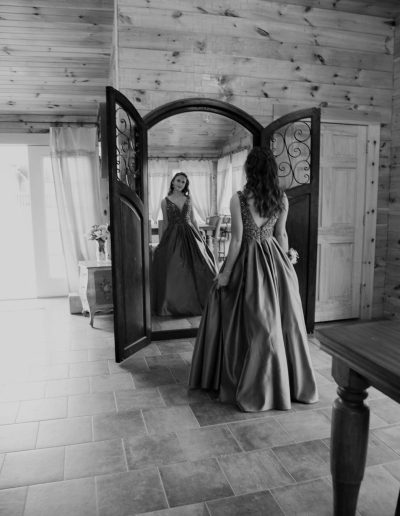 A bride in a satin wedding gown admires her reflection in an ornate full-length mirror inside a rustic wood-paneled venue.