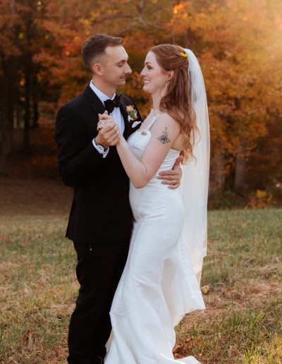 Bride and groom dancing together outdoors in formal wedding attire with autumn foliage in the background.