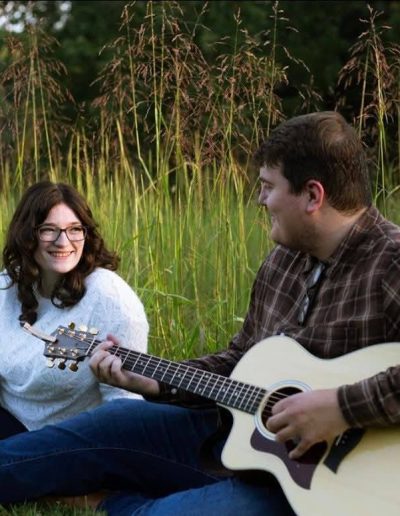 A young couple sitting in a grassy area, one playing guitar while the other smiles at them.