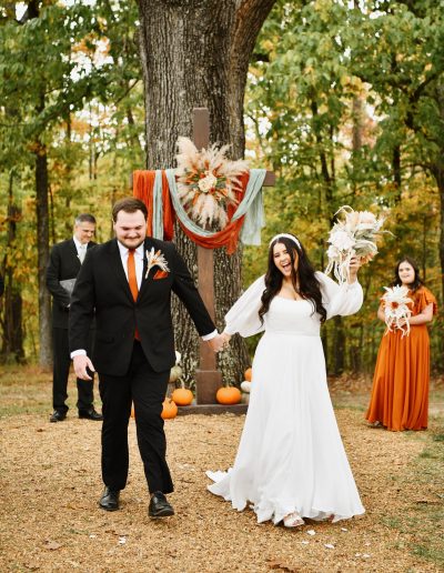 Bride and groom walking hand in hand after their wedding ceremony, surrounded by autumn foliage.
